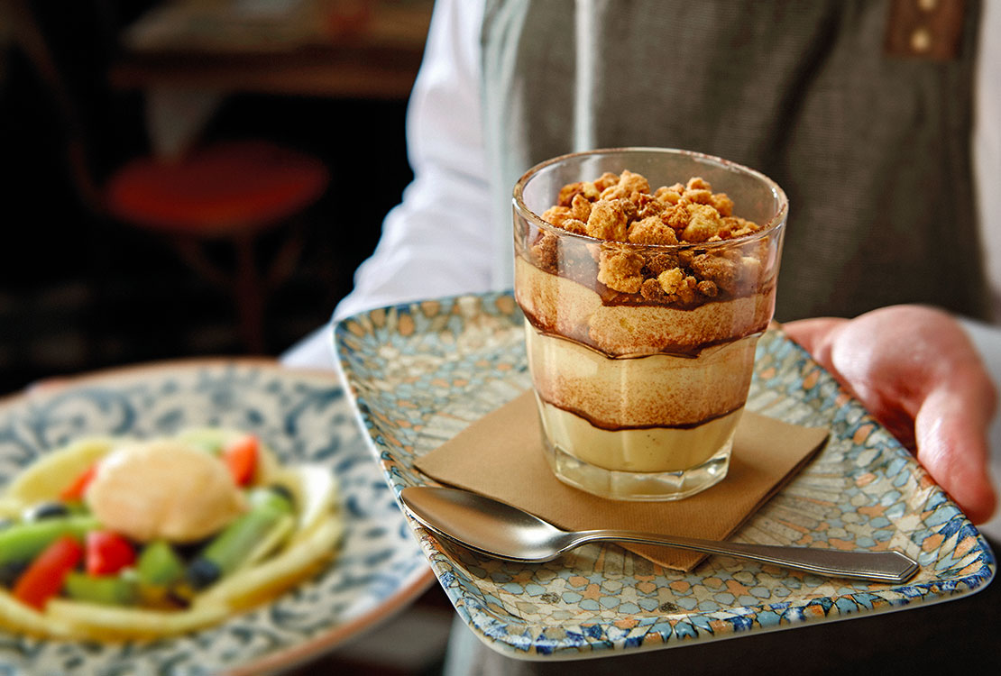 Un cameriere tiene in mano due dessert: un vassoio con un dolce al cucchiaio e un piatto con una macedonia con gelato.