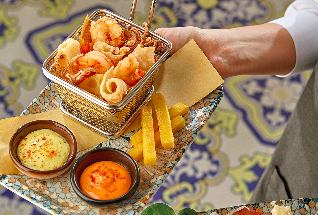 Two appetizers served on trays. The first appetizer is a fried fish with chips and sauces. The second appetizer is raw ham with cheeses.