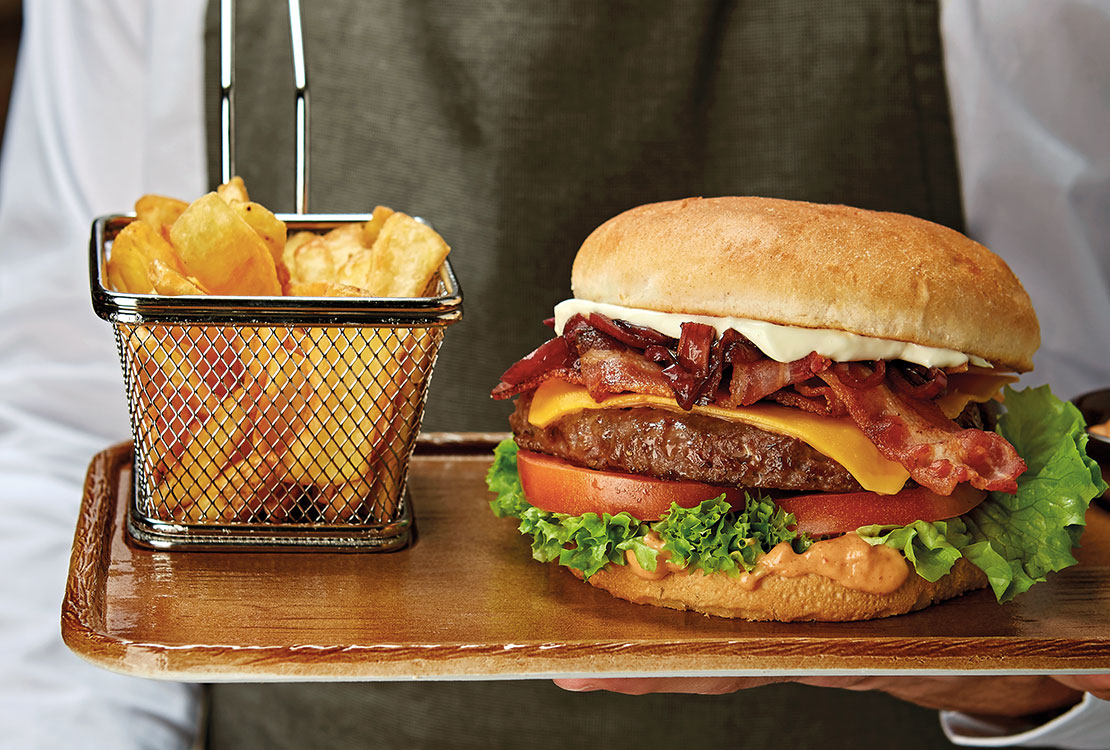 A waiter holds two main courses in his hand: a tray with hamburger and chips and a plate with tagliata and chips.