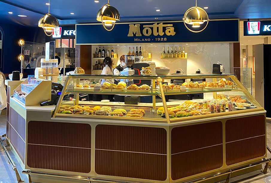 Interno del bar Motta Milano 1928 con il bancone e la vetrina contenente panini e croissant.