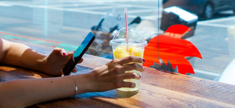 Utente seduto al bancone con 'vista parcheggio', con smartphone nella mano sinistra e bevanda nella mano destra.
